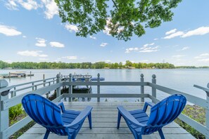 Une terrasse en bois avec deux chaises bleues donnant sur un lac paisible, une jetée avec des bateaux, et un ciel dégagé.
