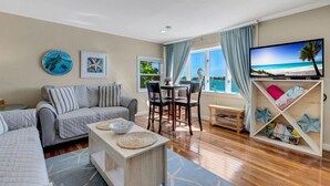 A living room with a sofa, a coffee table, and a TV on a stand. There is a window with curtains, a dining table with chairs, and a bookshelf. - 🌴 Bayview Beauty @ Coral Resort | Waterfront • Pool • Dock • Walk to Beach ➠ CR B5 (Clearwater Beach)