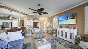 Een ruime woonkamer met een grote bank, een salontafel en een aan de muur bevestigde televisie. Er is een plafondventilator, een zithoek met stoelen, en een keuken op de achtergrond.
