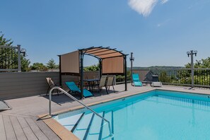 A swimming pool with a wooden deck, lounge chairs, a covered seating area, and a view of the landscape.