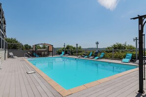 A rectangular swimming pool with a wooden deck, surrounded by blue lounge chairs and a covered seating area.