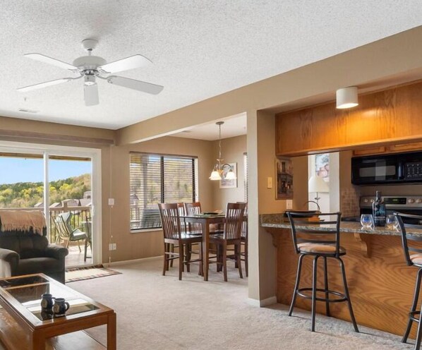 A spacious living area with a dining table, chairs, a coffee table, a ceiling fan, and a kitchen area with a microwave and bar stools.