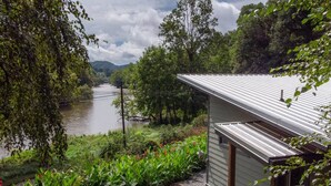 A house with a metal roof overlooking a river, surrounded by lush greenery and a garden with a chair. - Time Flies on the River (Bryson City)