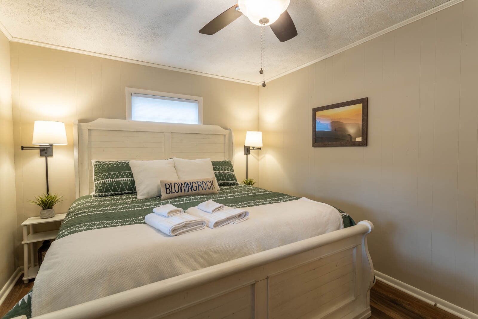 A neatly made bed with a pillow labeled "BLOWINGROCK," two bedside lamps, a ceiling fan, and a framed picture on the wall.