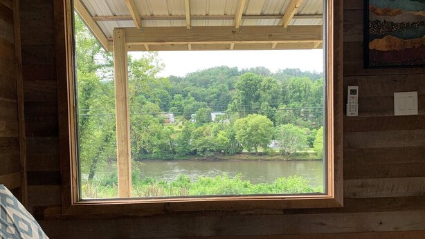 A bedroom with a large window overlooking a lake and forest. - Ship Shak on the River (Bryson City)