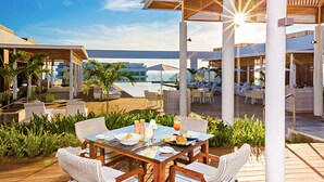 Un área de comedor al aire libre con vistas a la piscina y al océano, que cuenta con mobiliario de ratán blanco y una mesa de madera.