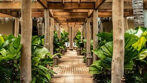 Un paseo techado con pilares de madera y techo de paja, bordeado por exuberante vegetación tropical.