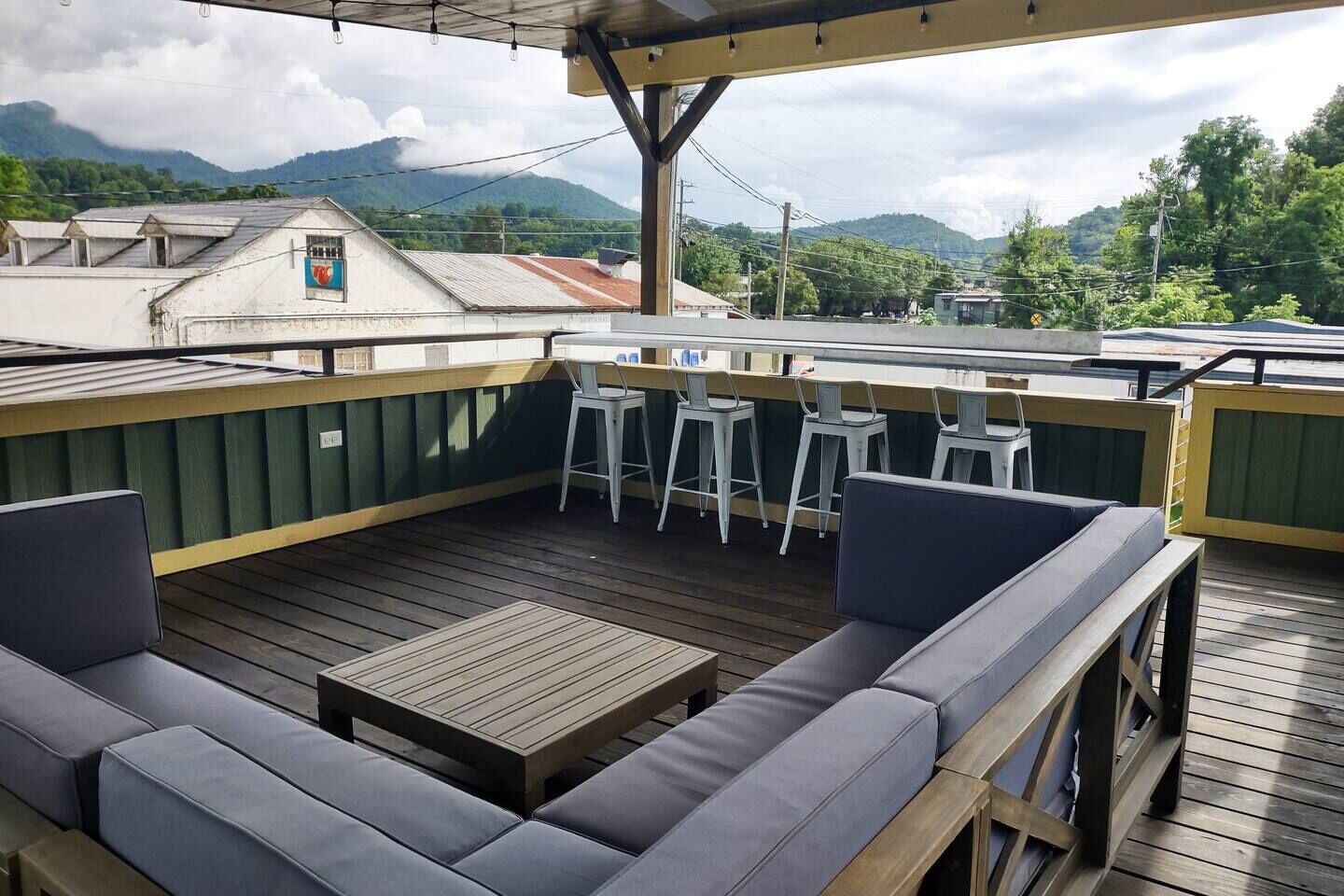 A covered outdoor seating area with wooden flooring, cushioned benches, and bar stools.