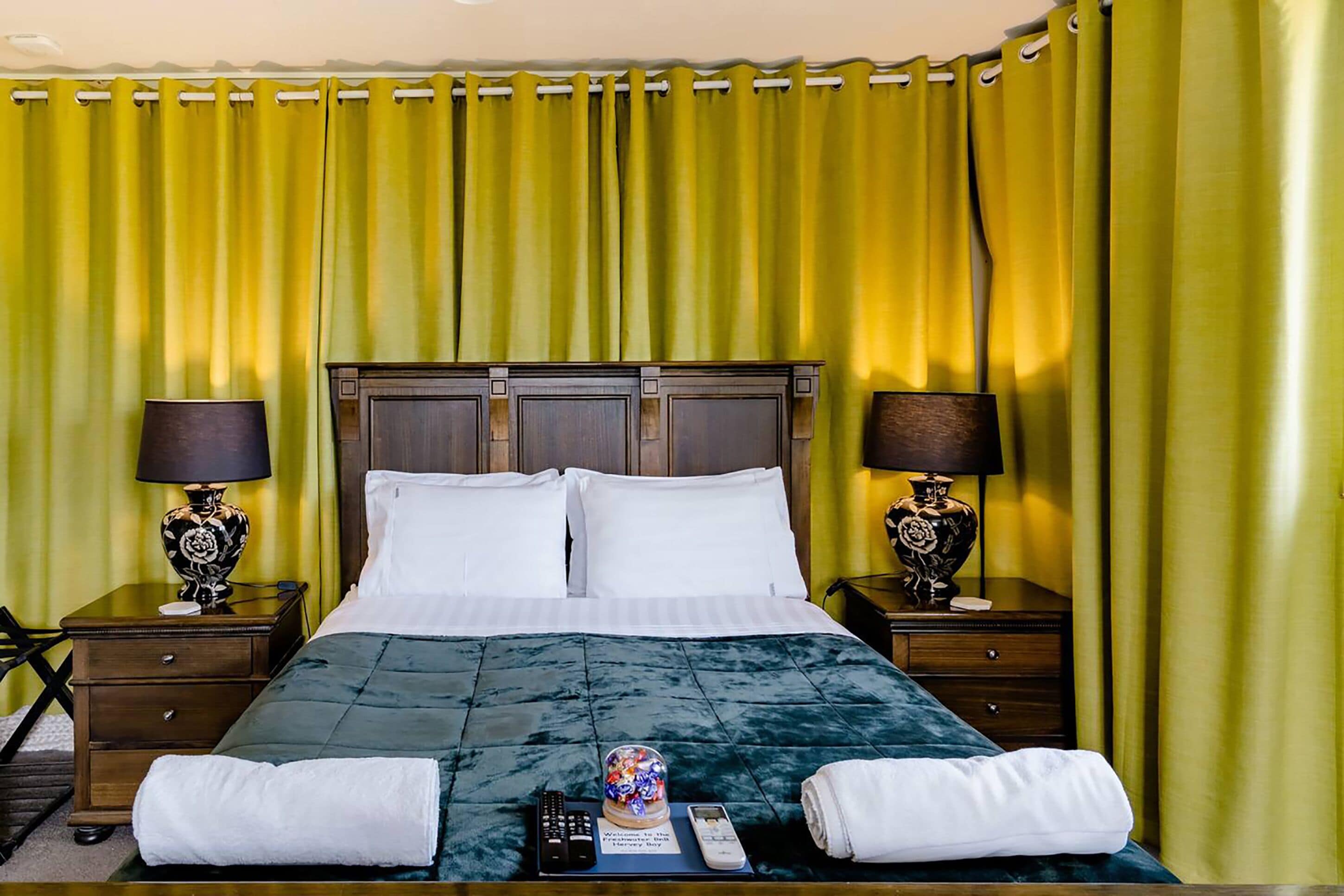 A hotel room with a large bed, two bedside tables, and a green curtain backdrop.