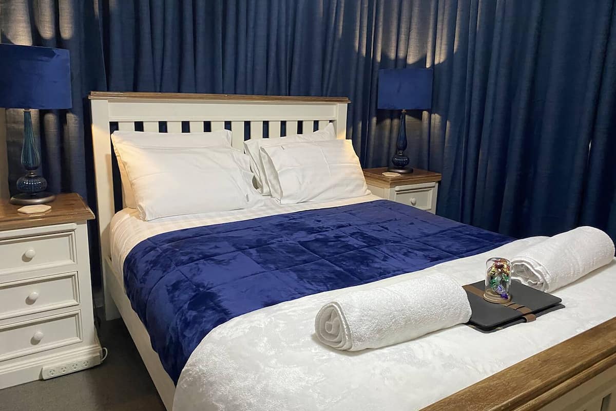 A neatly made bed with a blue bedspread and white pillows, a wooden headboard, and a white nightstand with a lamp.