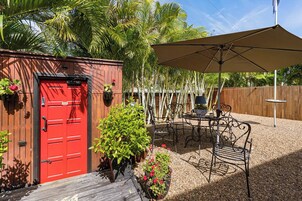 Una puerta roja con una valla de madera, un patio con sillas y una mesa de metal, y un paraguas grande.