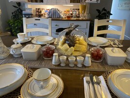 Desayuno con una mesa preparada para cuatro personas, incluyendo platos, tazas y una bandeja de frutas.