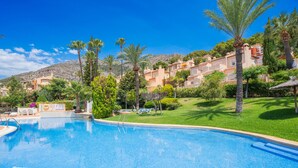 A swimming pool with a clear blue water, surrounded by palm trees and a building with multiple balconies. - CostaBlancaDreams - Dinamarca in Altea Hills (Altea)