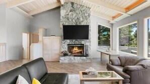 A modern living room with a stone fireplace, a flat-screen TV mounted above it, and a large window providing a view of the outdoors. - Sea Ranch Ocean Views 4 bedrooms + office (Sea Ranch)