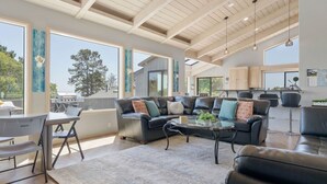 A spacious living area with a large couch, coffee table, and dining table. There are large windows offering views of the outdoors. - Sea Ranch Ocean Views 4 bedrooms + office (Sea Ranch)
