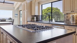 A modern kitchen with a large island, wooden cabinets, a black countertop, and a view of trees through a large window. - Sea Ranch Ocean Views 4 bedrooms + office (Sea Ranch)