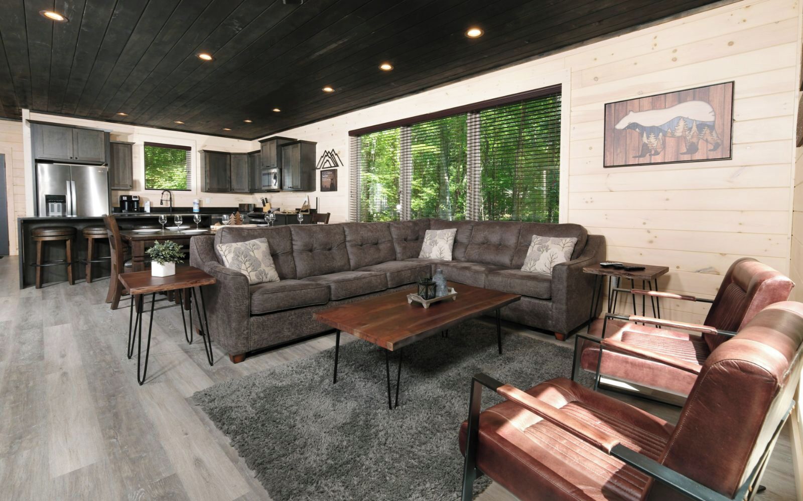 Ein modernes Wohnzimmer mit einem Ecksofa, einem Couchtisch und Ledersesseln. Im Hintergrund ist eine Küche mit dunklen Holzschränken und Edelstahlgeräten zu sehen.