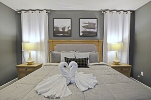 Un lit bien fait, avec deux serviettes disposées en forme de cygne, une tête de lit en bois, deux lampes de chevet et deux tableaux encadrés accrochés au mur.
