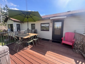 A wooden deck with a table and chairs, a red Adirondack chair, a green umbrella, and a house with a door and windows. - Mahnomen Cottage: Hike, Bike, Fish, & Relax only 2 hrs north of the Twin Cities! (Deerwood)