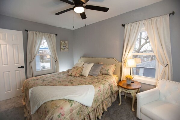 Un dormitorio con una cama, ventilador de techo, mesita de noche con lámpara y una silla.