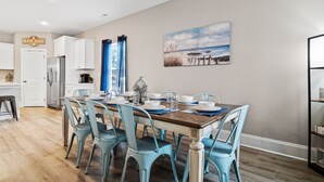 A dining area with a wooden table and blue chairs, a kitchen with white cabinets, and a beach-themed painting on the wall.