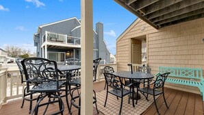 Un patio techado al aire libre con mesas y sillas de metal, una valla de madera y un edificio con balcón al fondo.