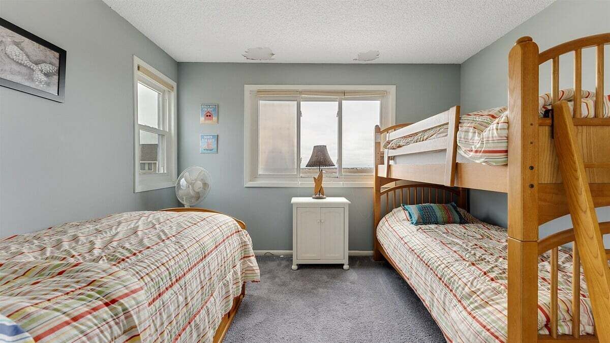 Une chambre avec deux lits superposés, une table de chevet avec une lampe, un petit meuble et une fenêtre avec des stores.