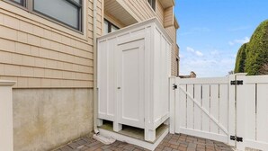 Una caseta utilitaria blanca al lado de una casa con revestimiento color beige y cerca blanca.