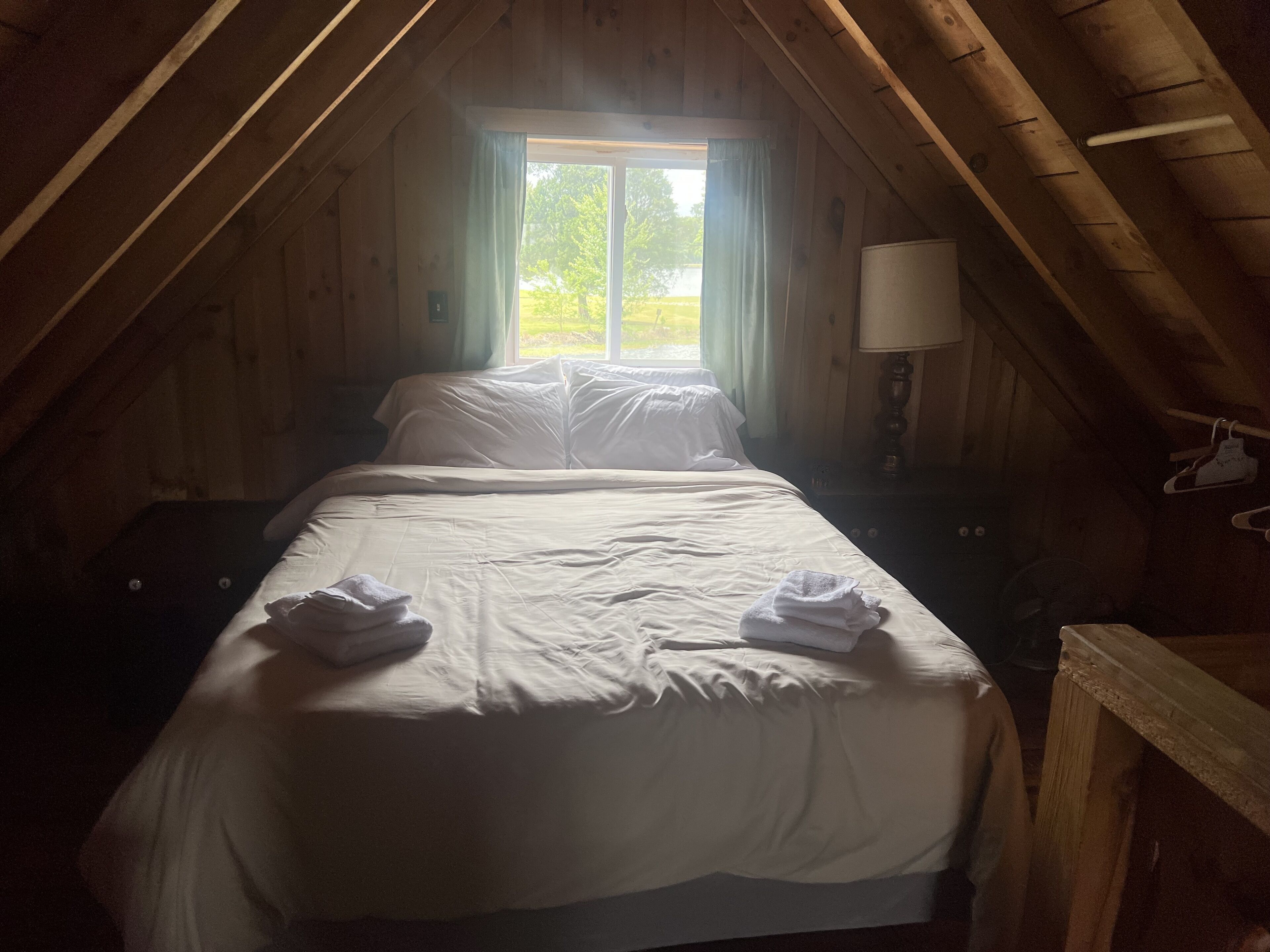 A single bed in a wooden attic room with a window, a lamp, and a wooden chair.