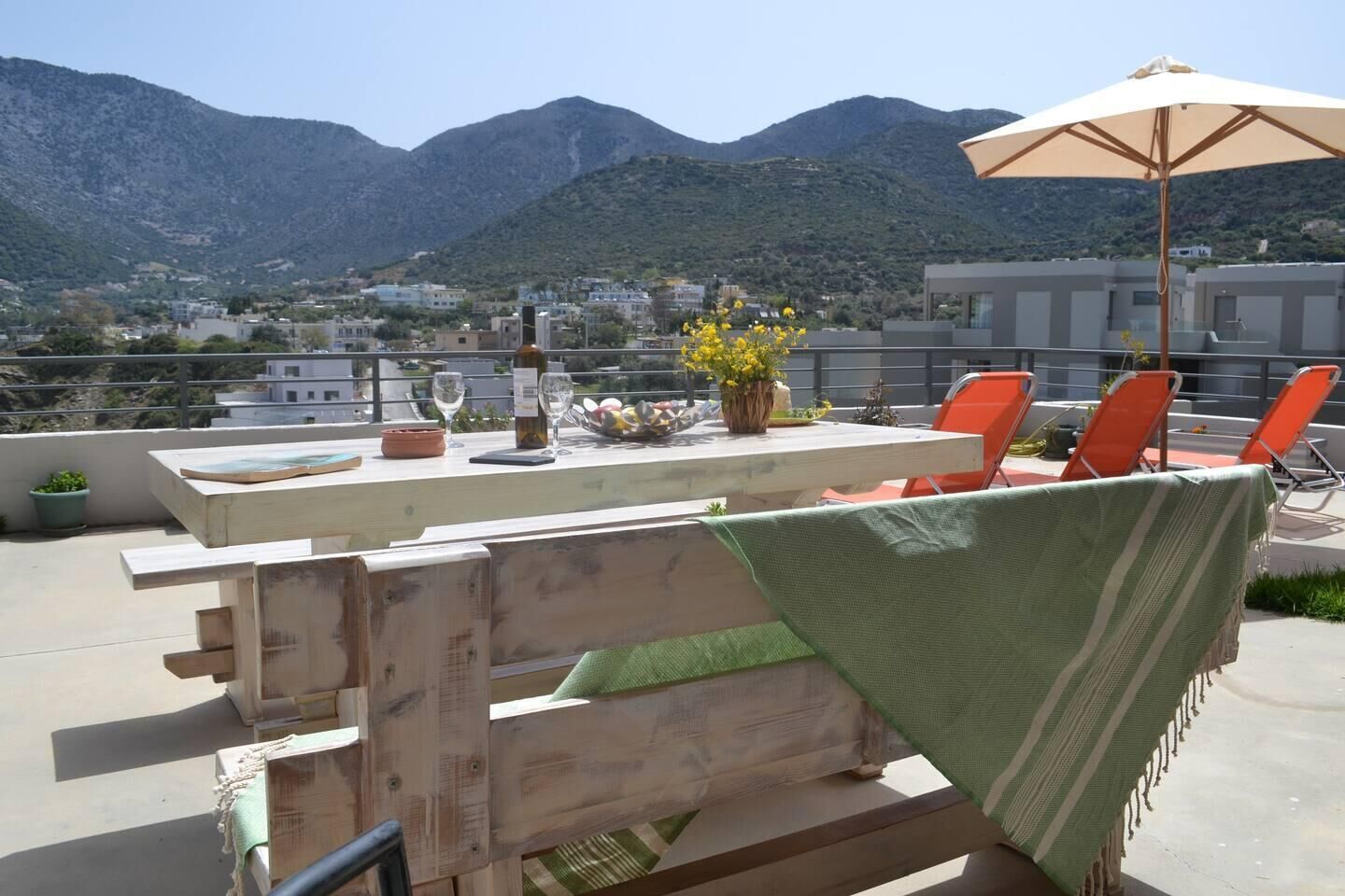 A rooftop terrace with a wooden table, orange chairs, a green towel, and a large umbrella.