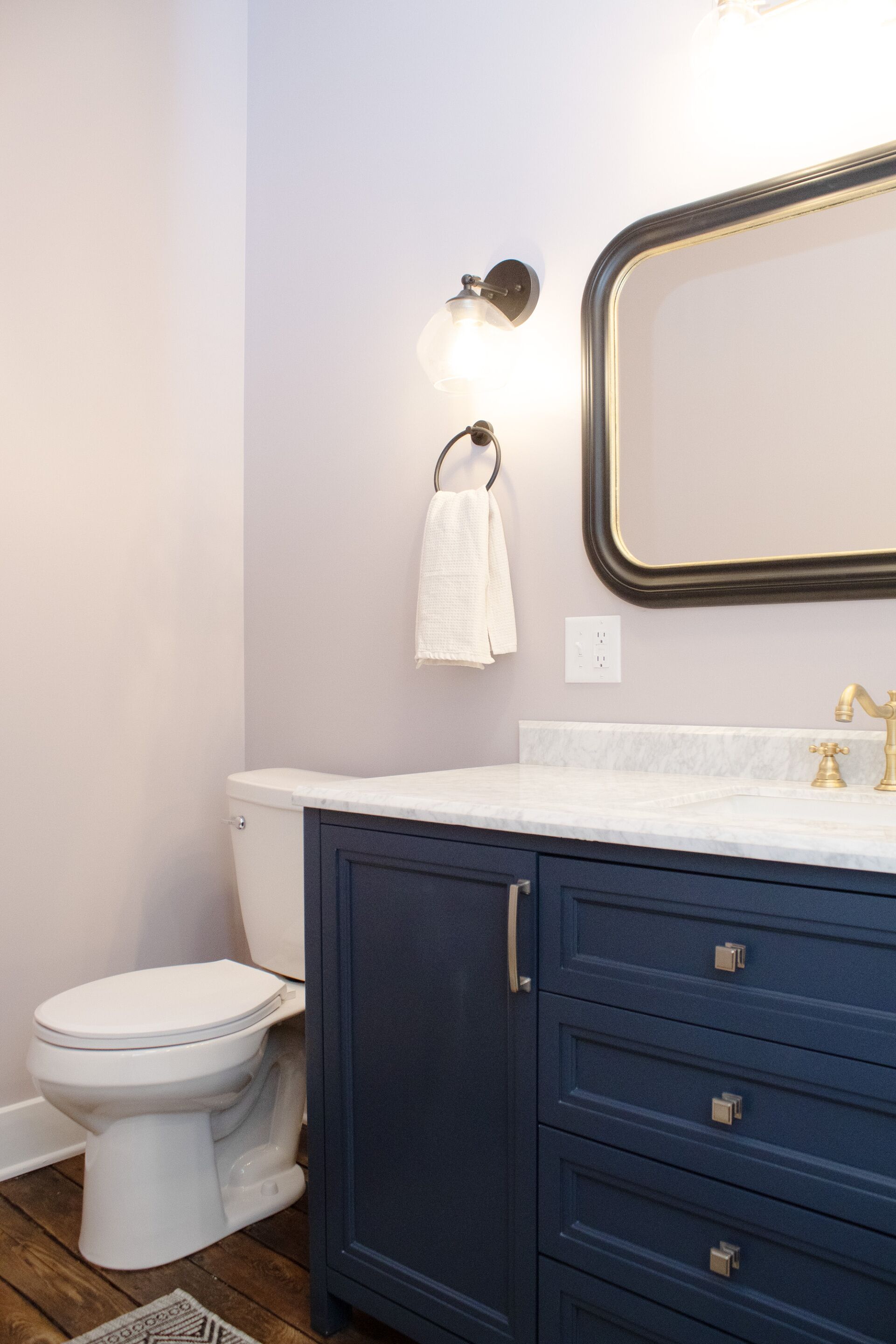 A bathroom with a white toilet, a blue vanity with drawers, a gold faucet, and a wall-mounted mirror.
