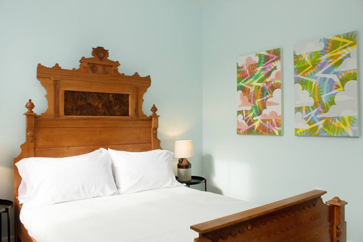 Cama bem arrumada com cabeceira de madeira, mesa de cabeceira com abajur e duas pinturas abstratas na parede.