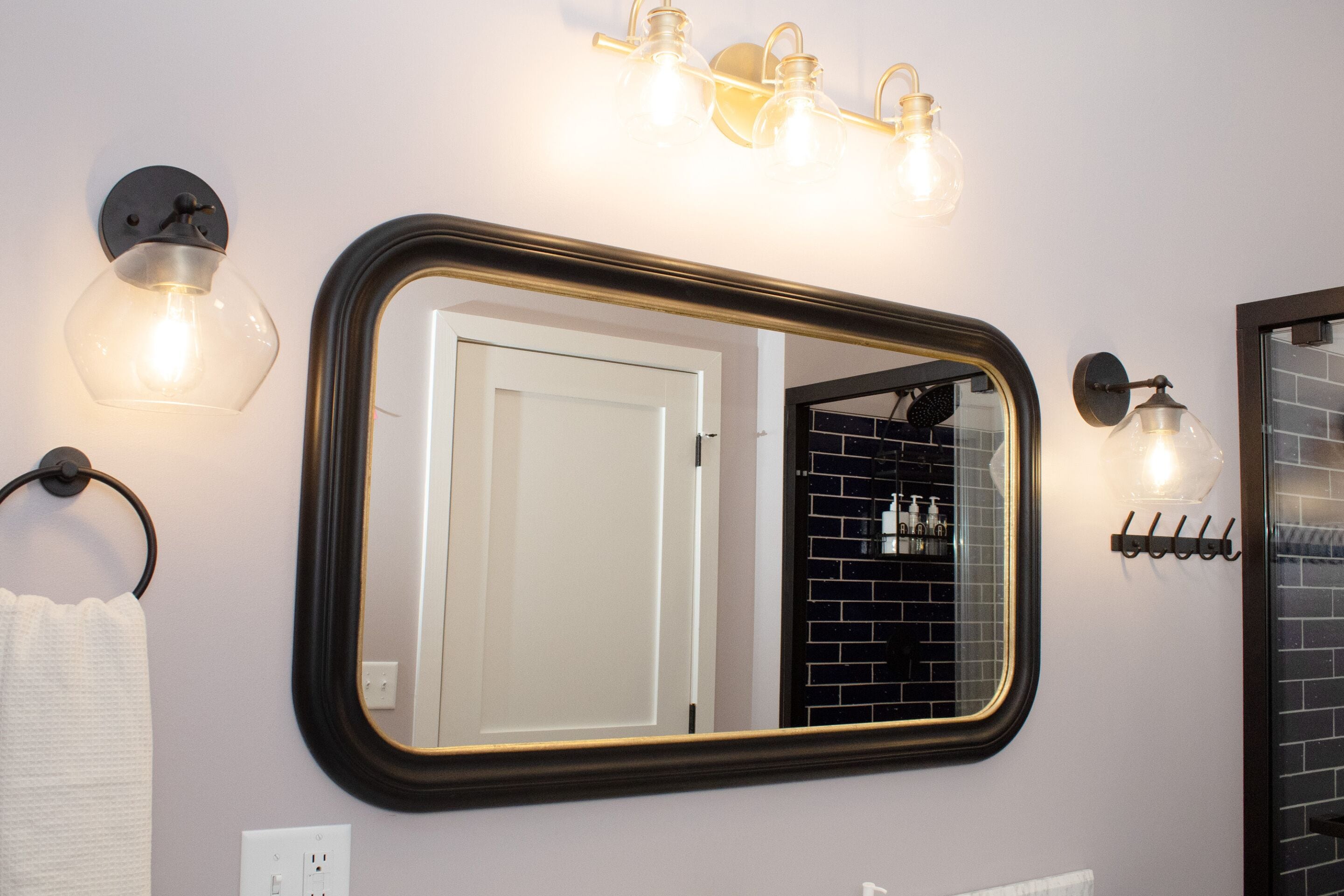 A bathroom with a large mirror, wall-mounted light fixtures, a towel rack, and a door.
