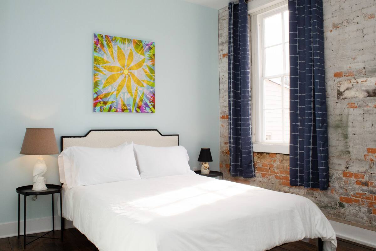 Quarto com cama, mesa de cabeceira, luminária, pintura na parede e janela com cortinas.