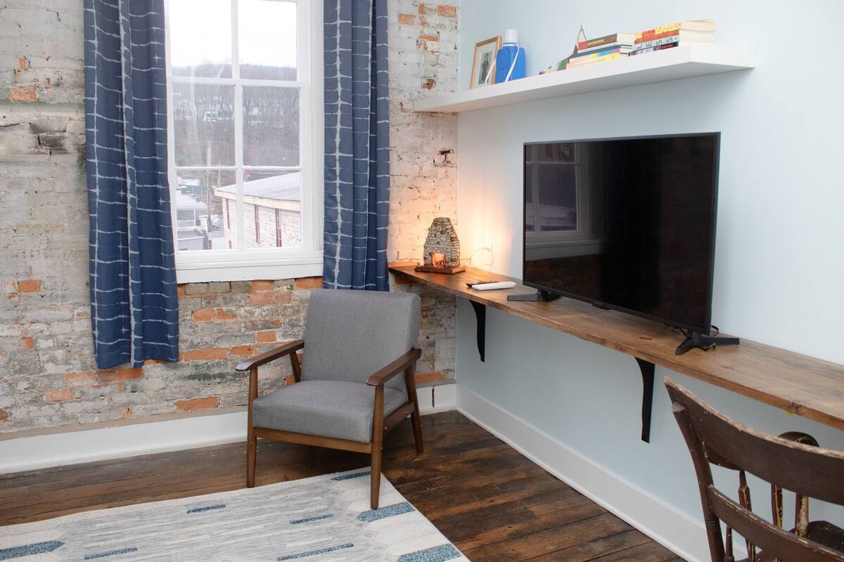Um quarto aconchegante com parede de tijolos, uma janela grande com cortinas, uma cadeira de madeira, uma TV em uma prateleira e uma pequena mesa com abajur.