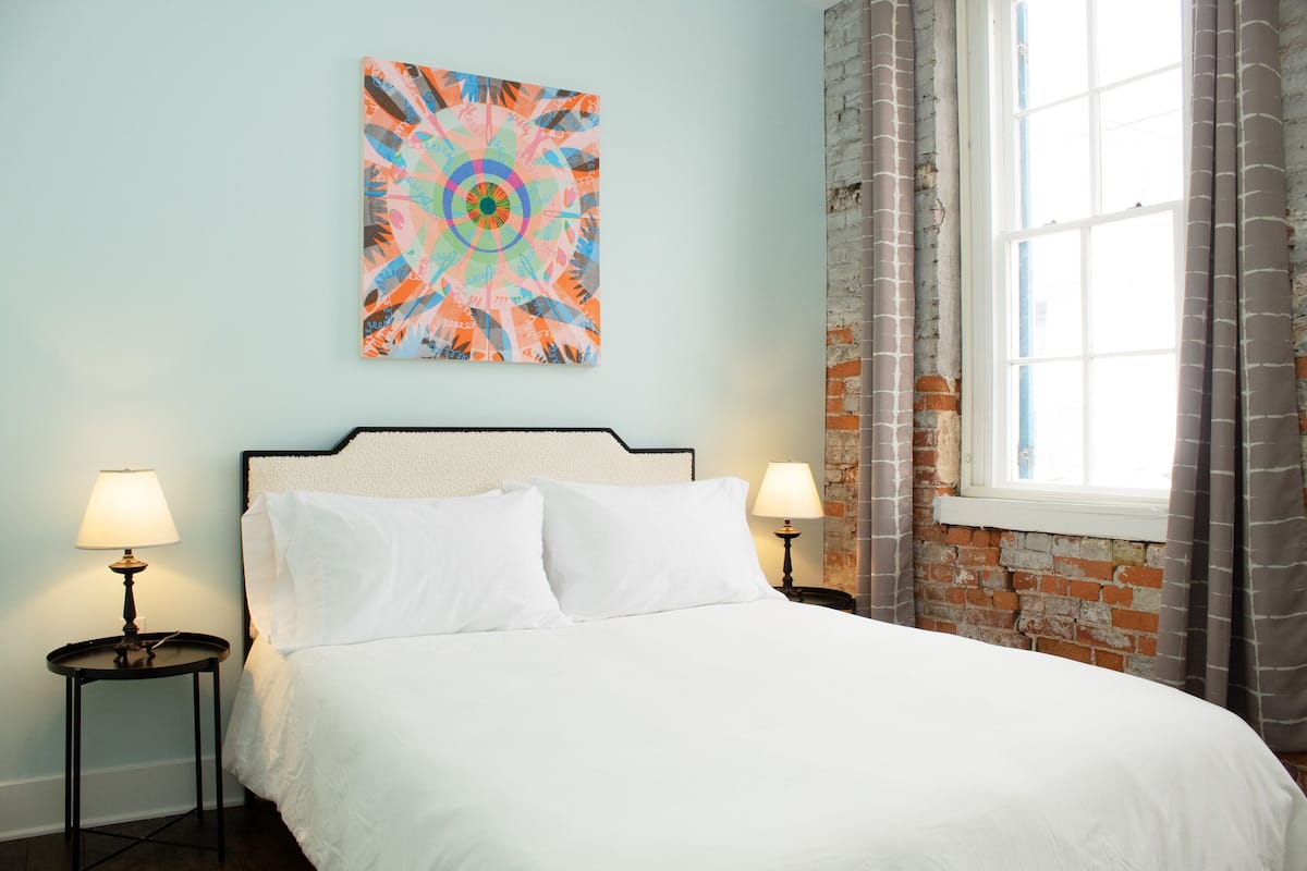 Cama bem arrumada com lençóis brancos em quarto com paredes azul claro, uma pintura abstrata colorida acima da cama e uma janela com cortinas.
