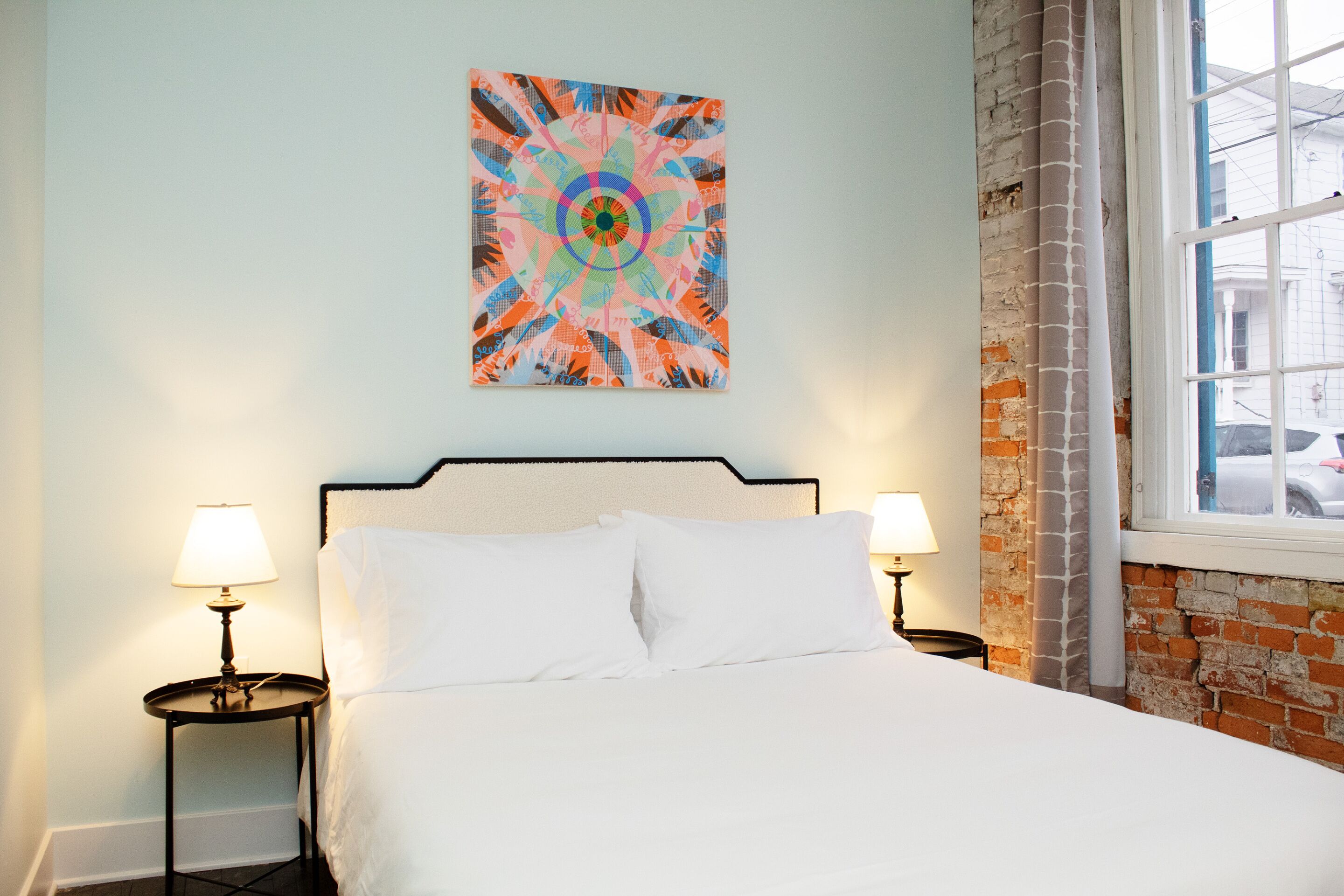 Habitación de hotel con cama, dos lámparas de noche, un cuadro abstracto de colores en la pared y ventana con vistas a la calle.