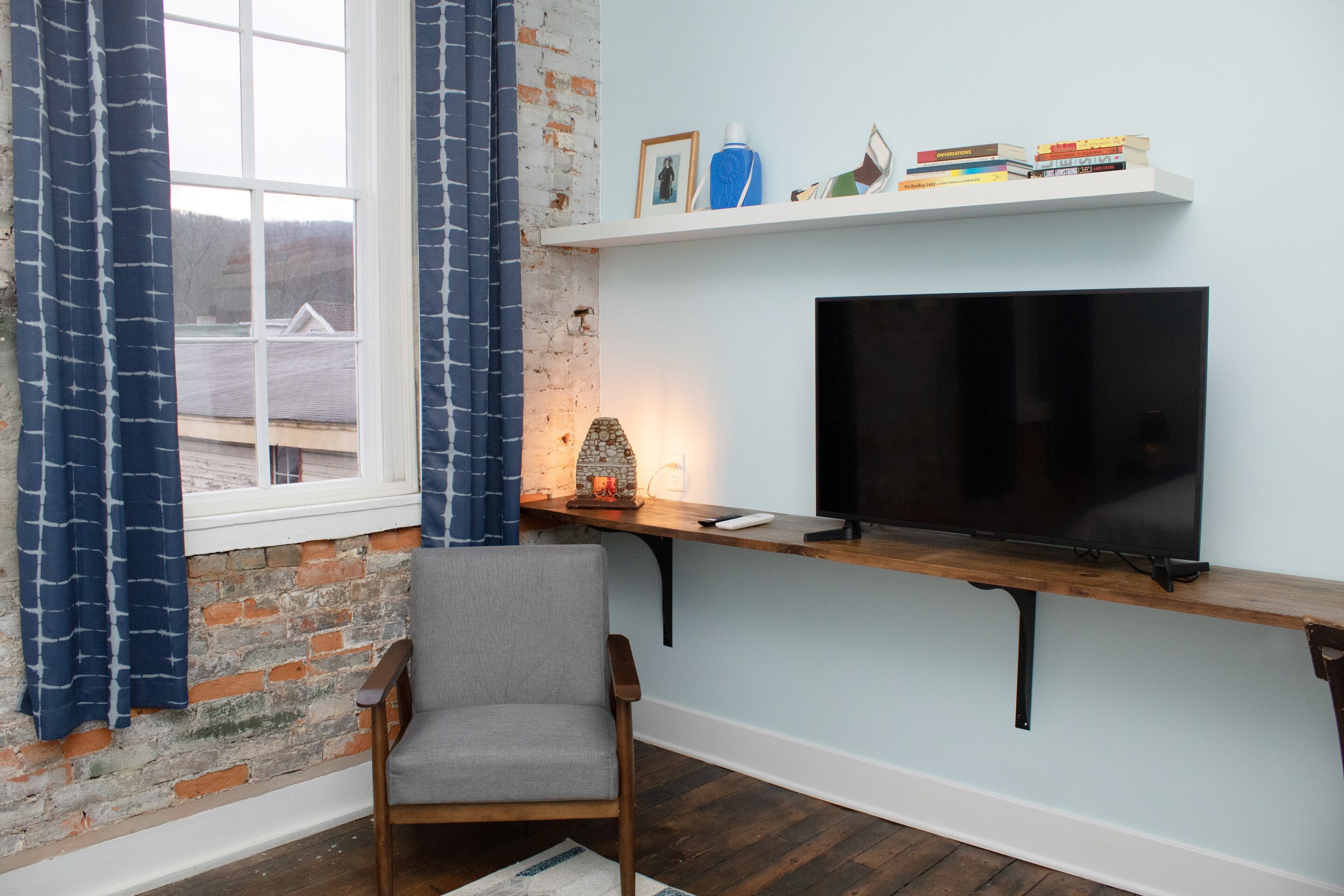 Una habitación con pared de ladrillo, una ventana con cortinas azules, una silla de madera, un televisor sobre una estantería y una mesita con objetos decorativos.