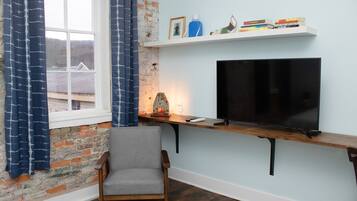 Una habitación con pared de ladrillo, una ventana con cortinas azules, una silla de madera, un televisor sobre una estantería y una mesita con objetos decorativos.