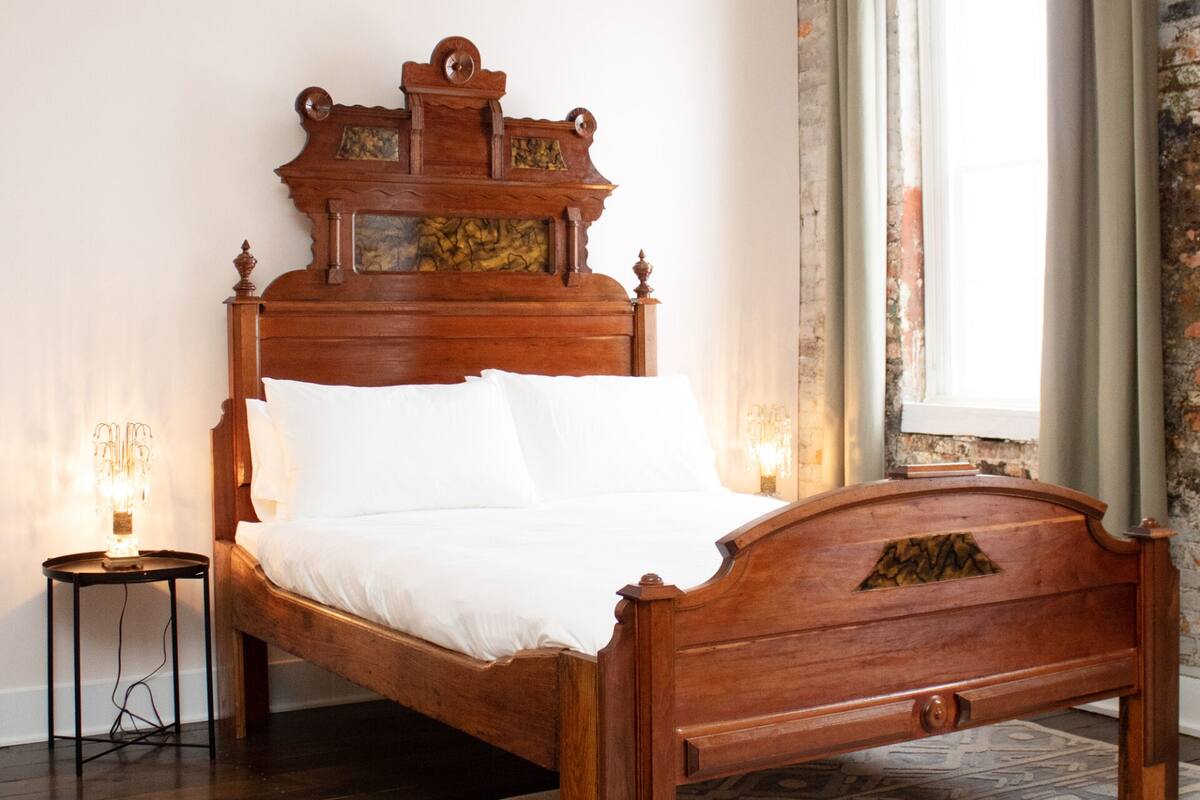 Uma cama de madeira com cabeceira, uma mesa de cabeceira com abajur e um tapete estampado sobre piso de madeira.