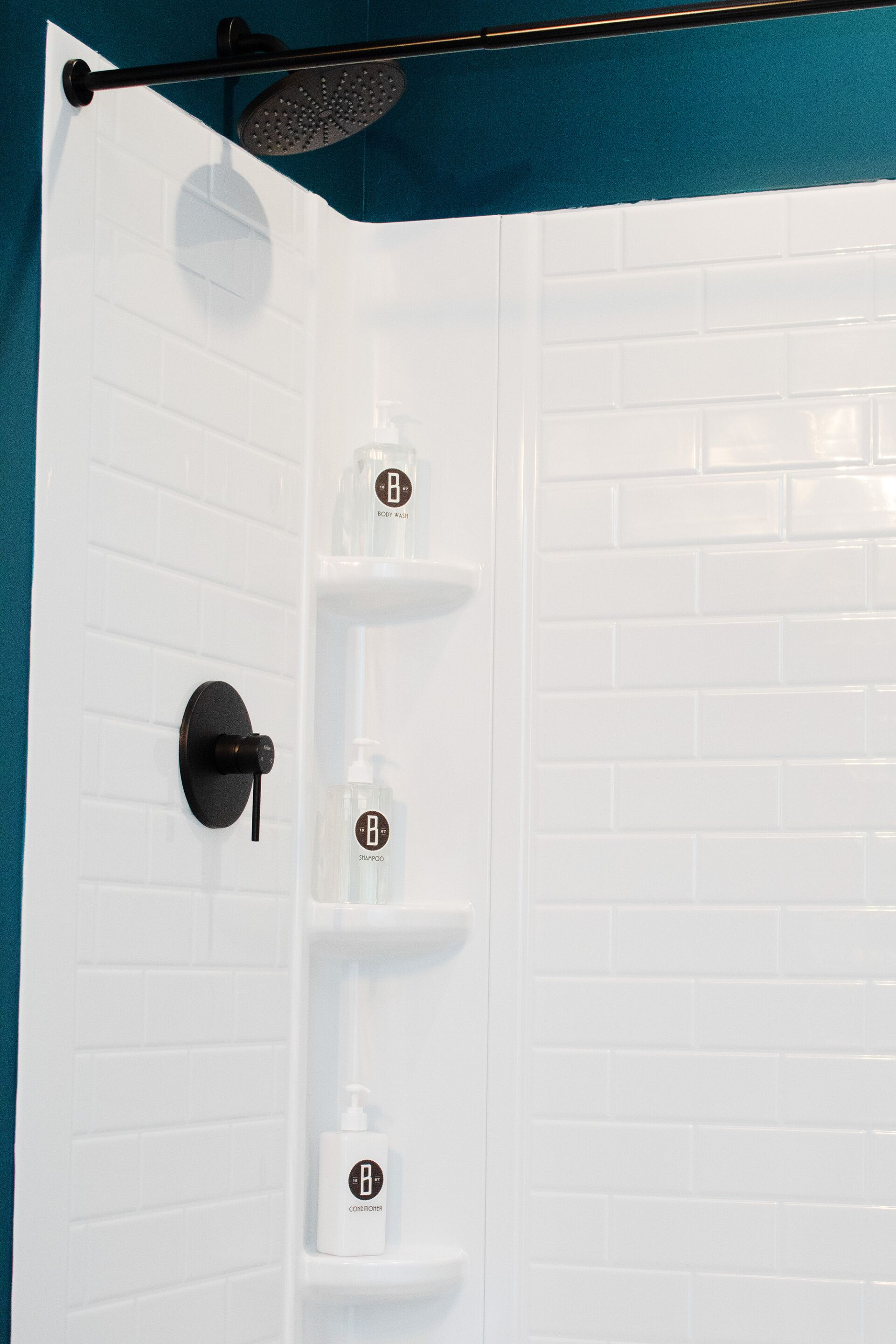 Zona de ducha con pared de azulejos blancos, ducha negra y tres repisas con botellas etiquetadas 'BODY WASH', 'SHAMPOO' y 'CONDITIONER'.