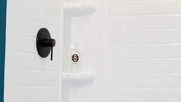 Zona de ducha con pared de azulejos blancos, ducha negra y tres repisas con botellas etiquetadas 'BODY WASH', 'SHAMPOO' y 'CONDITIONER'.