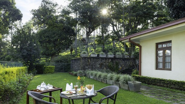 An outdoor dining area with a stone wall, a small building with a window, and a well-maintained lawn. - Olive Bungalow - Jetwing Select (Welimada)