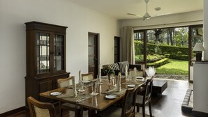 A dining area with a wooden table set for six, a dark wooden cabinet, and a view of a well-manicured garden through large windows. - Olive Bungalow - Jetwing Select (Welimada)