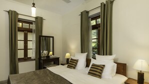 A neatly made bed with a patterned bedspread, a bedside table with a lamp, and a window with curtains. - Olive Bungalow - Jetwing Select (Welimada)