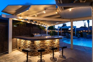An outdoor bar area with a stone wall, bar stools, and a pool in the background. - Luxury Oasis, Hot Tub, Outdoor Kitchen and Grill, Arcade Room, Sleep up to 13 (Fresno)