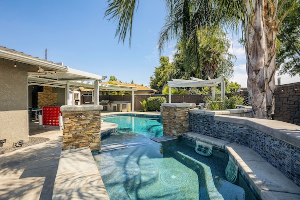 A swimming pool with a stone and tile border, a covered patio area, and a stone wall.