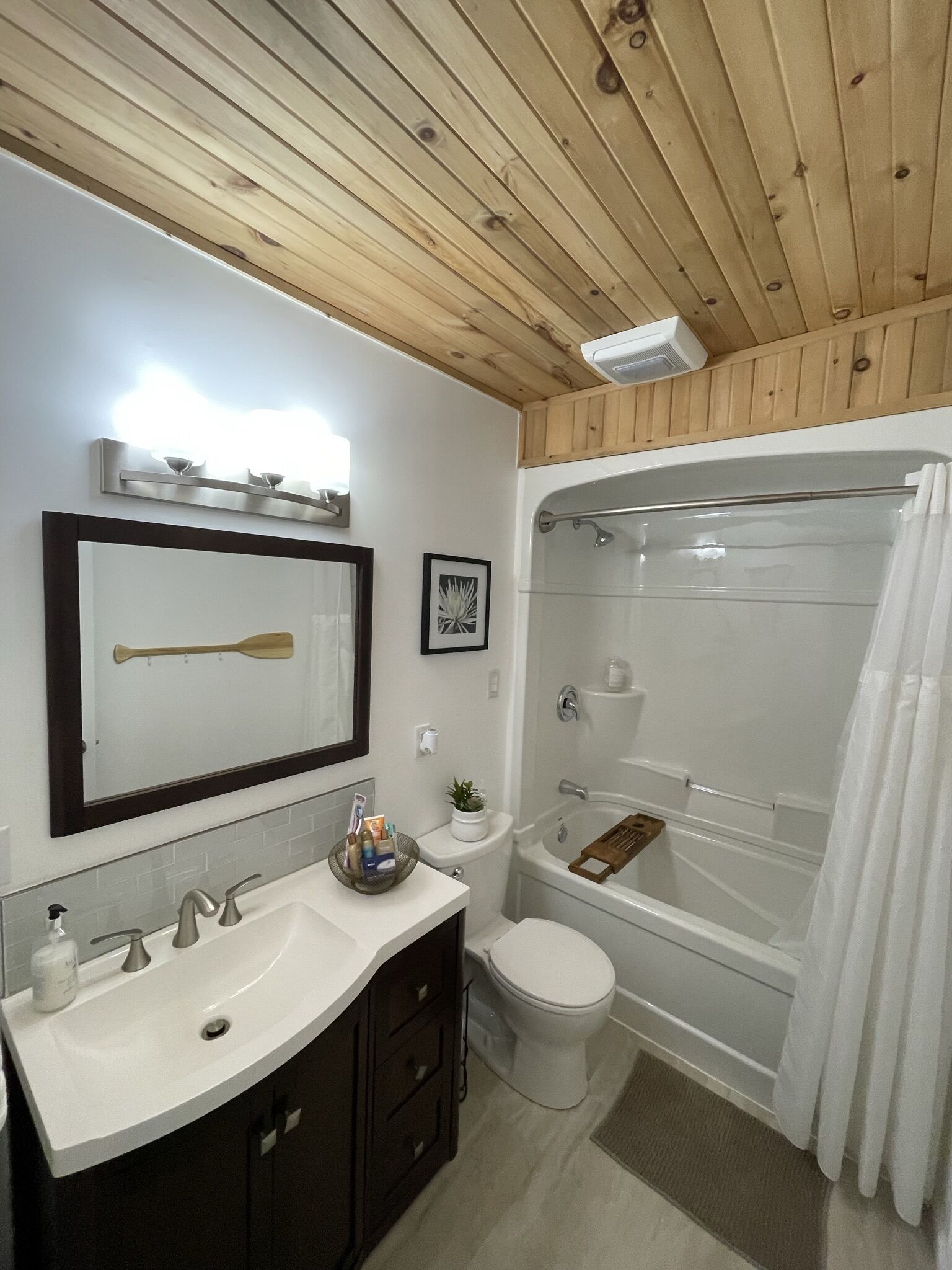 A bathroom with a wooden ceiling, a white sink with a mirror, a toilet, and a bathtub with a shower curtain.