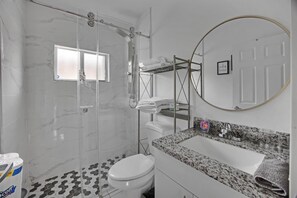A modern bathroom with a glass shower enclosure, a toilet, a sink with a granite countertop, and a mirror with a circular frame. - Close To Airport, Miami & Wynwood New Modern Home! (Miami)