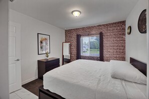 A bedroom with a brick wall, a bed, a dresser, a mirror, and a window with blinds. - Close To Airport, Miami & Wynwood New Modern Home! (Miami)